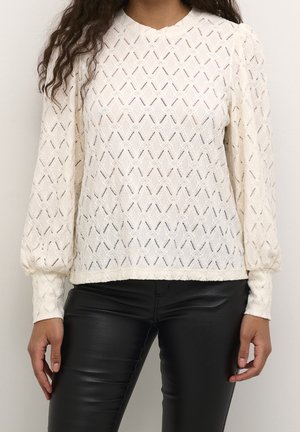 Vrouw draagt een crèmekleurige blouse met lange mouwen, textuur en diamantpatronen, gecombineerd met strakke zwarte leren broek, staand tegen een effen achtergrond.