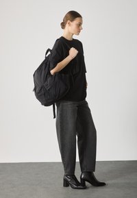 Svart ryggsäck i tyg med flera fickor, bärs över en svart oversized T-shirt, tillsammans med högmidjade grå jeans och svarta klackskor.