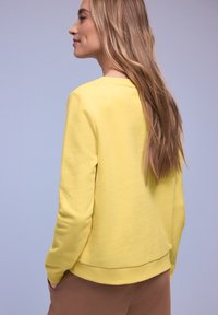 Femme aux longs cheveux blonds portant un sweat-shirt jaune et un pantalon marron, vue de dos sur un fond violet clair.