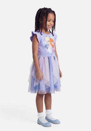 Niña joven con el cabello trenzado, vestida con un vestido morado temático de Frozen, con mangas con volantes y falda de tul en capas, calcetas blancas y zapatos azules.