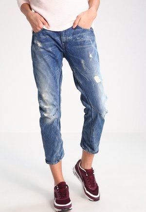 Vaqueros desgastados de color azul claro con un corte relajado, que presentan desvanecidos, bordes deshilachados y detalles de costura visibles. Llevados con zapatillas de deporte marrón burdeos.