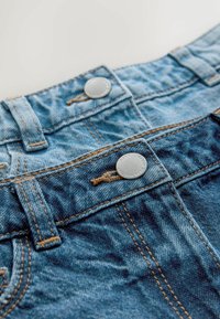 Šviesiai mėlyni džinsiniai šortai su dviem sidabro spalvos metaliniais mygtukais, kontrastingomis rudos spalvos siūlėmis ir tekstūruota medžiaga su subtiliu išblukimu.