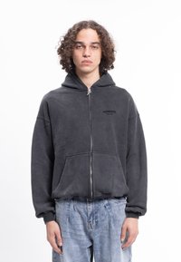 Černá mikina na zip z měkké látky s klokaní kapsou. Má kapuci a nenápadné logo na hrudi. Nositelná s světle modrými džínami.