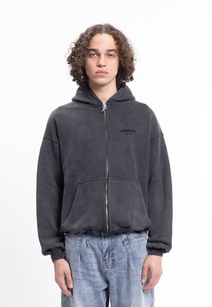 Sudadera negra con cremallera hecha de tela suave con un bolsillo canguro. Cuenta con capucha y un logo sutil en el pecho. Combinada con vaqueros azul claro.