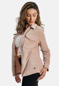 Giacca camicia in corduroy rosa chiaro con due tasche frontali, chiusure a bottoni e orlo curvo, caratterizzata da una leggera texture e un dettaglio con logo sul lato.
