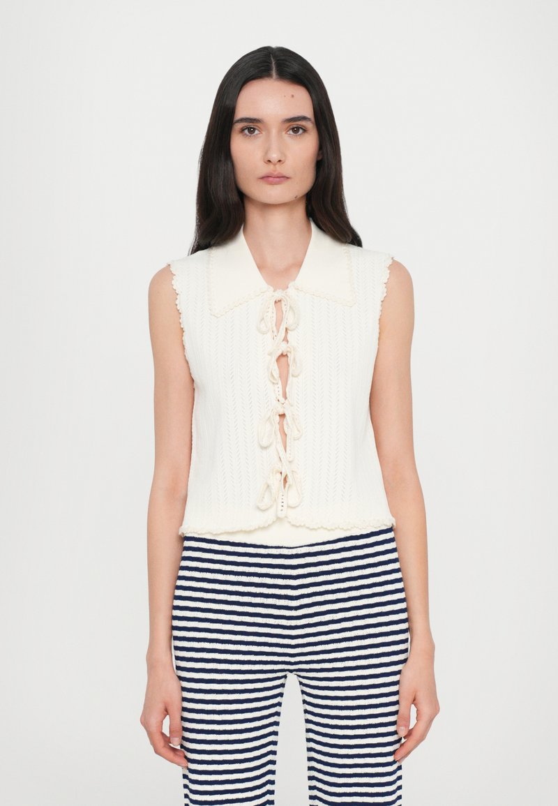 Débardeur en tricot crème avec un col à volants, doté d'une fermeture à nouer à l'avant et d'un motif texturé. Associé à un pantalon rayé bleu et blanc.