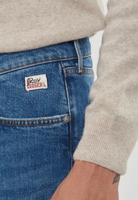 Jeans in denim blu con etichetta "Roy Rogers" vicino alla tasca, indossati con un maglione lavorato a maglia beige con maniche e un tatuaggio visibile sul polso.
