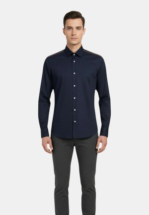 Uomo che indossa una camicia aderente blu marina scuro con sottili strisce sulle spalle e pantaloni grigi, in piedi contro uno sfondo bianco semplice.