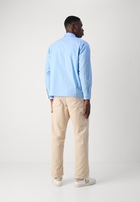 Lichtblauw geruit overhemd met lange mouwen, beige cargo broek en witte sneakers, staand op een effen achtergrond.