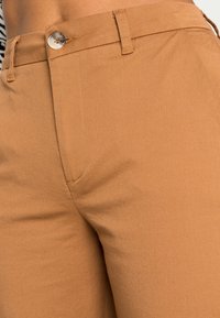 Pantaloni beige con una texture liscia, dotati di passanti per cintura e chiusura a bottone. Il tessuto sembra resistente con una vestibilità sagomata.