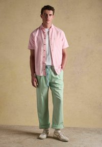 Jeune homme debout à l'intérieur, portant une chemise à manches courtes rose clair boutonnée par-dessus un t-shirt blanc, un pantalon rayé vert et des baskets beige.
