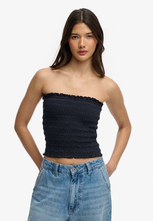 Femme portant un crop top noir texturé sans bretelles et un jean bleu taille haute, les mains dans les poches, devant un fond uni.