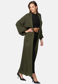 Lång olivgrön stickad cardigan med ribbad textur, vida ärmar och en framficka, i kombination med en svart kortärmad topp och eleganta svarta byxor.