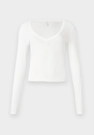 Balta, garām piedurknēm, V-veida kakla pieguļošs īss tops ar vienkāršu, gludu auduma tekstūru neitrālā fonā.