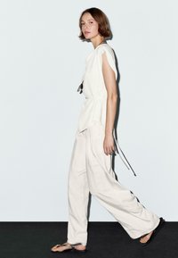 Top sans manches blanc au fit décontracté, noué à la taille ; associé à un pantalon ample de couleur claire ; modèle portant des sandales noires.