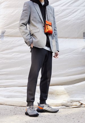 Personne portant un blazer à rayures grises sur un hoodie noir, un pantalon de jogging noir, des baskets grises avec des accents orange, et un petit sac bandoulière orange.