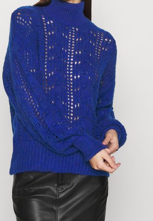Femme portant un pull en tricot bleu à motif ajouré et manches longues, associé à une jupe en cuir noire.