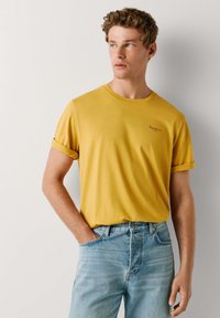 Žlté bavlnené tričko s rolovanými krátkymi rukávmi, okrúhlym výstrihom a vyšitým logom na hrudi, v kombinácii so svetlomodrými rifľami.