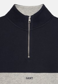 Quarter-zip pull i marineblå bomull med et lys grått stoffpanel og "GANT"-logo. Har sølvglidelås og ribbet krage.