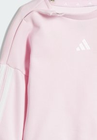 Svetloružová mikina s bielymi akcentmi a textúrovaným golierom; má malé biele logo na hrudi a dve biele prúžky na rukávoch.