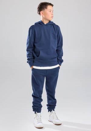Marineblauwe hoodie met een voorzak, gecombineerd met bijpassende joggers en gemusterde witte sneakers. Gemaakt van zacht materiaal.