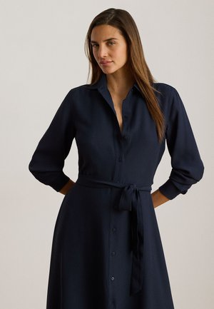 Vrouw met lang bruin haar, die een marineblauwe jurk met knopen draagt en een gestrikte tailleband, tegen een effen lichte achtergrond.