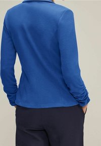 Blå fleecjacka med skräddarsydd passform, långa ärmar med rynkdetalj och krage. Texturerad tyg, lämplig för avslappnad klädsel.