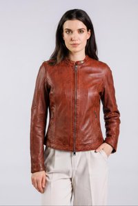 Pitkähiuksinen nainen, jolla on päällään ruskea nahkatakki, jossa on vetoketju, ja vaaleanbeiget housut, seisoo käsi taskussa yksinkertaisen taustan edessä.