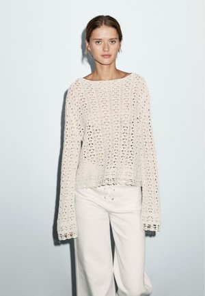 Light cream, knitted sweater with a loose fit, lengthy sleeves, open weave pattern, and scalloped hem. Paired with white, drawstring pants.