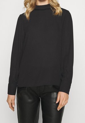 Vrouw draagt een zwarte blouse met lange mouwen en een met kralen versierde kraag en een zwarte leren broek, staand tegen een effen achtergrond.