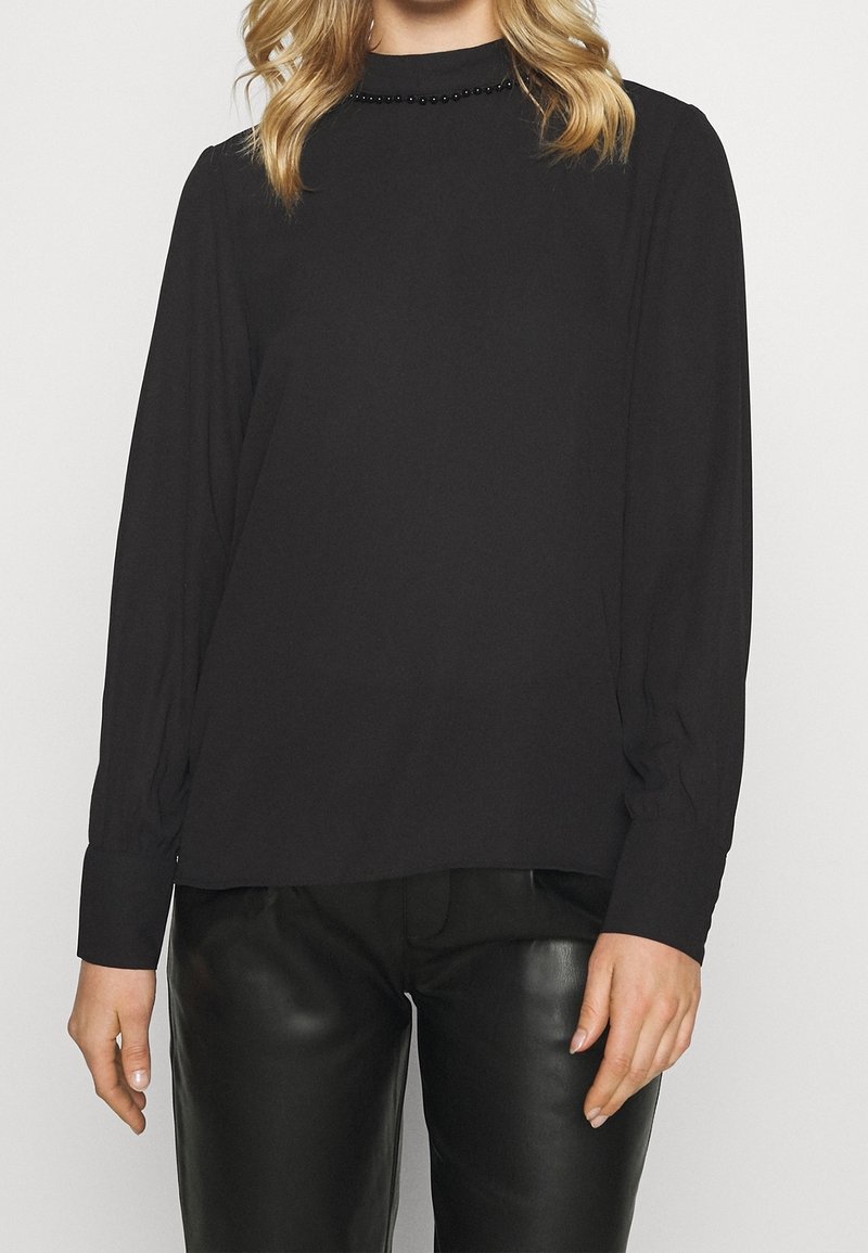 Femme portant un chemisier noir à manches longues avec un col orné de perles et un pantalon en cuir noir, debout devant un fond uni.