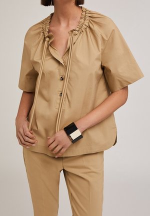 Blusa beige de manga corta con escote fruncido y cordones, combinada con pantalones beige y una pulsera gruesa en blanco y negro.