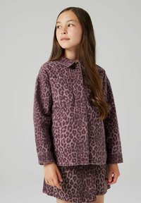 Jauna mergaitė su ilgais rudais plaukais, apsirengusi violetiniu leopardų rašto švarku ir jam derančia sijonėliu, žiūri šiek tiek į kairę.