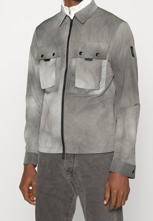 Homme portant une veste grise zippée avec deux poches poitrine à boutons et un pantalon gris assorti sur un fond uni.
