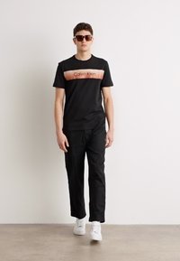 Svart bomull t-skjorte med kort erme og en kontrastfylt lys rosa horisontal logo-panel som leser "Calvin Klein." Kombinert med svarte bukser og hvite joggesko.