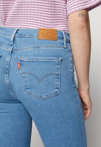 Ljusblå denimjeans med hög midja, med en brun läderlapp på baksidan och en signatur röd etikett på fickan. Texturerad tyg.