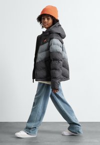 Puffytakki mustana ja harmaana, jossa on huppu ja oranssi logo. Yhdistettynä vaaleansinisillä leveälahkeisilla farkuilla ja valkoisilla tennareilla.