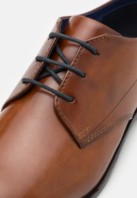 Brauner Lederschuh mit glatter Oberfläche, schwarzen Schnürsenkeln, runden Zehen und dezenten Nähten an der Seite.