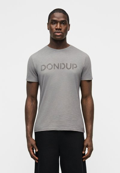 Homme debout de face portant une chemise grise à manches courtes avec le texte "DONDUP" et un pantalon noir, sur un fond uni.