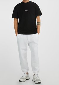 Černé bavlněné tričko s potiskem "YPS" v bílé barvě, kombinované s bílými tepláky a šedými sneakerami s černými akcenty a texturovanou podrážkou.