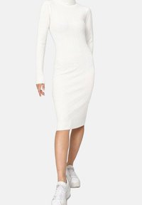 Robe tricotée blanche à côtes avec des manches longues et un col haut, ajustée jusqu'au mollet, portée avec des baskets blanches. Texture lisse.