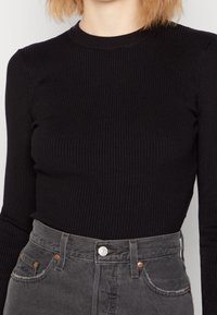Schwarzes, geripptes Langarmoberteil kombiniert mit hochgeschnittenen grauen Jeans, die über Fronttaschen und Metallknopfakzenten verfügen.