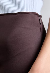 Fustă maro cu o textură netedă, dotată cu un fermoar pe lateral. Materialul pare ușor lucios, în contrast cu un top bleu deschis deasupra.