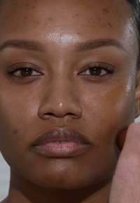 Nahaufnahme eines Gesichts, das Make-up aufträgt. Merkmale sind ein glatter, hellbrauner Teint, ausgeprägte Augenbrauen und volle Lippen.