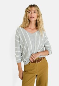 Mujer con cabello rubio ondulado que lleva un suéter de rayas grises y blancas de cuello en V y pantalones mostaza con cinturón marrón, mirando hacia un lado.