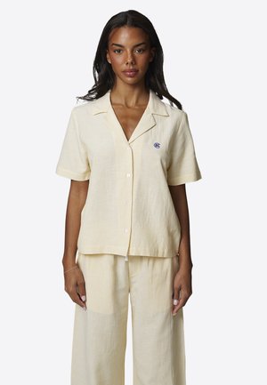 Kobieta w jasnżółtej koszuli zapinanej na guziki z krótkimi rękawami w cienkie prążki oraz pasujących szerokich spodniach, stojąca na jednolitym tle.