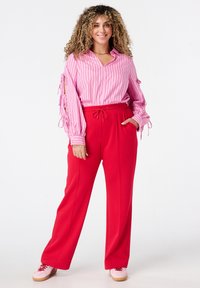 Glunderende vrouw met krullend haar, gekleed in een roze gestreepte blouse met vastgebonden mouwen, rode joggingbroek met aansnoerkoord en roze sneakers, staand met haar handen in haar zakken.
