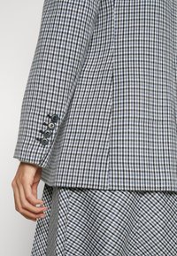 Blazer a cuadros en azul marino y blanco, con tres botones negros en el puño y una tela estructurada con una textura suave.