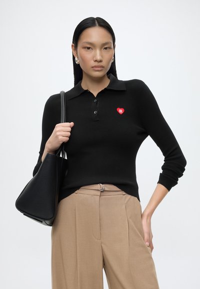 Femme portant un polo noir à manches longues avec un logo cœur rouge, pantalon beige, tenant un sac à main noir sur l'épaule.