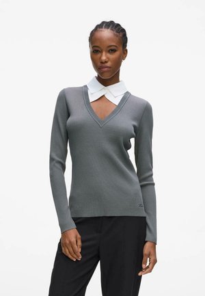 Femme aux cheveux tressés portant un pull gris côtelé à col en V sur un col blanc, associé à un pantalon noir, debout devant un fond blanc uni.